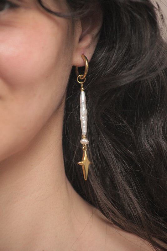 Mujer con pendientes artesanales de acero inoxidable. Cada par presenta una perla natural auténtica acompañada de un delicado charm de estrella dorada, creando un diseño versátil que captura la luz con sofisticación. 